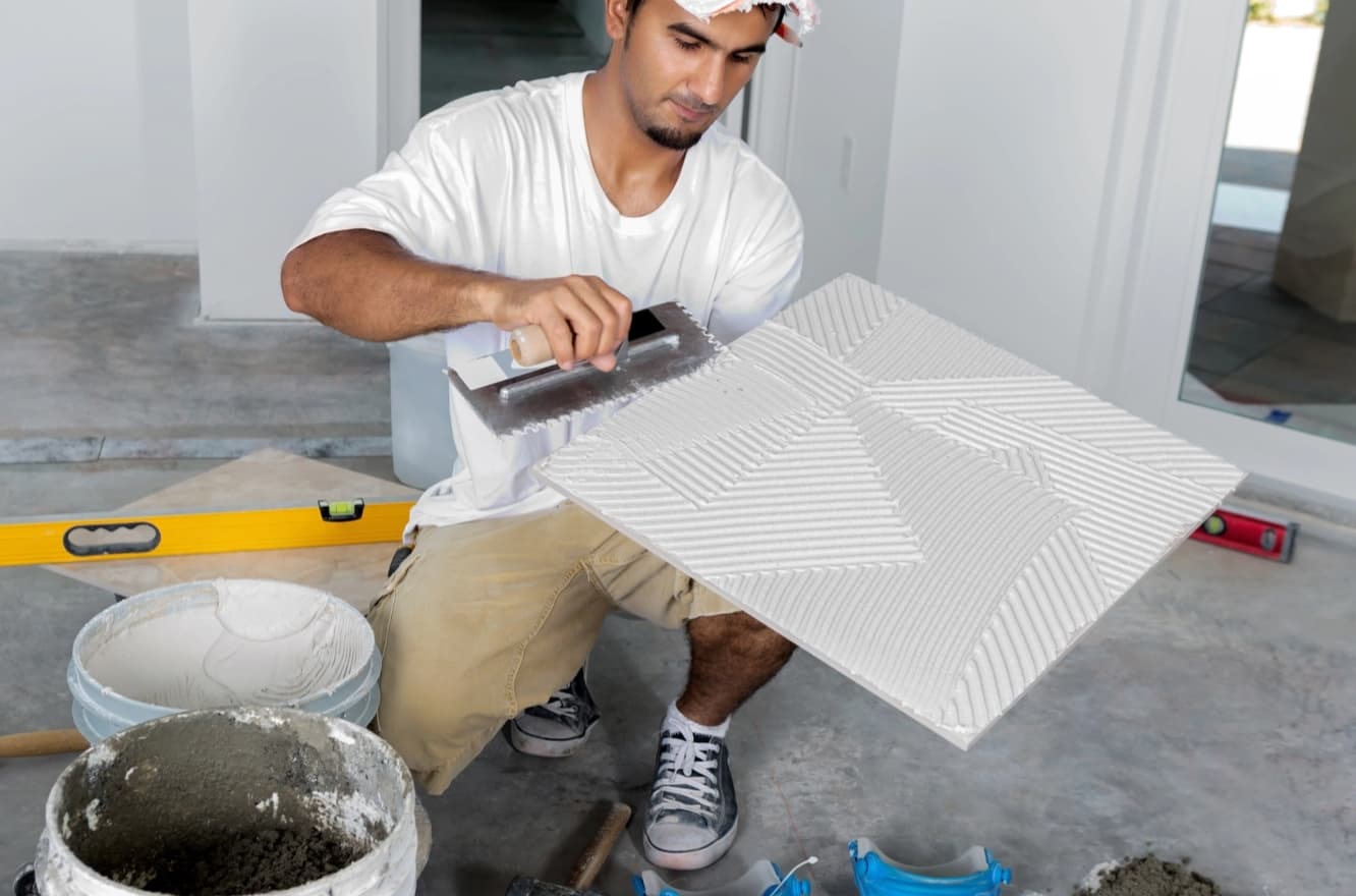 Focused tradesman applying adhesive with a notched trowel on a tile, surrounded by construction tools.